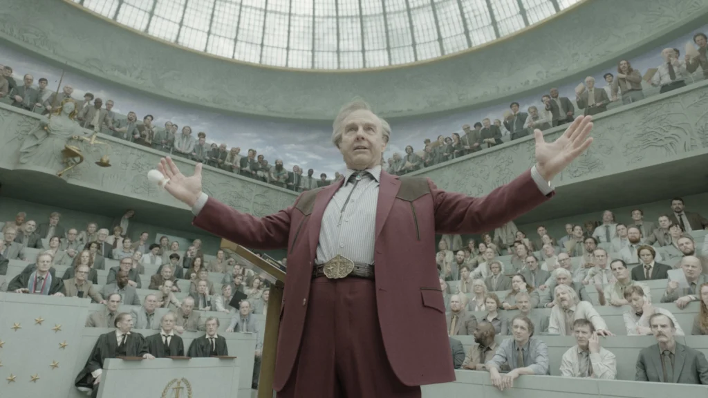 Tom Cruise as Digger Rockwell, in a maroon cowboy suit, extending his arms during a fiery monologue. He stands in a large, concrete room filled with drab people looking on in grey-colored clothing.